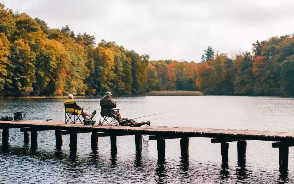 Tranquil image: Two individuals sitting on a lakeside dock, surrounded by serene waters and nature. A moment of relaxation by the lake's edge